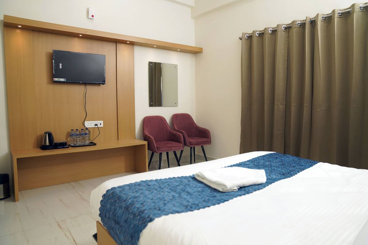 Habitación de hotel con cama, televisión montada en la pared, dos sillas, cafetera y espejo.
