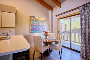 A dining area with a table, chairs, a vase of flowers, a painting on the wall, and a view through a glass door. - Front St Leavenworth Condo + Parking (Leavenworth)