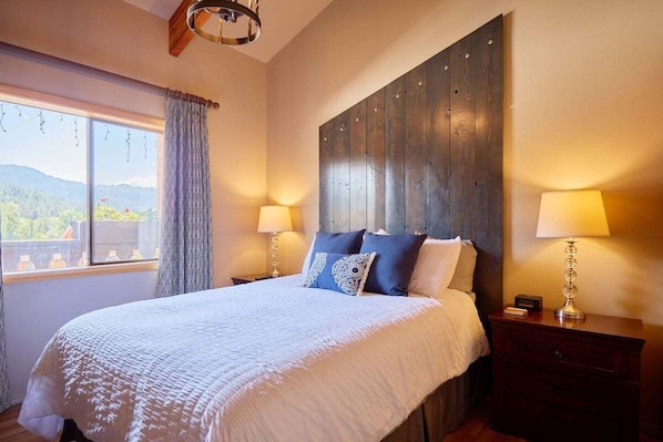 A bedroom with a large bed, two bedside lamps, a wooden headboard, a window with curtains, and a view of the outdoors. - Mountain Town Loft • Near Eats, Shops, All the Fun (Leavenworth)