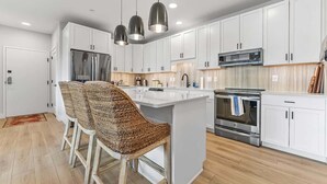 A modern kitchen with white cabinets, a central island with bar stools, stainless steel appliances, and pendant lighting. - Modern Condo Near DT TC | Beach + Bikes (Traverse City)