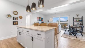 A modern kitchen with white cabinets, a central island, pendant lighting, and a dining area with wicker furniture. - Modern Condo Near DT TC | Beach + Bikes (Traverse City)