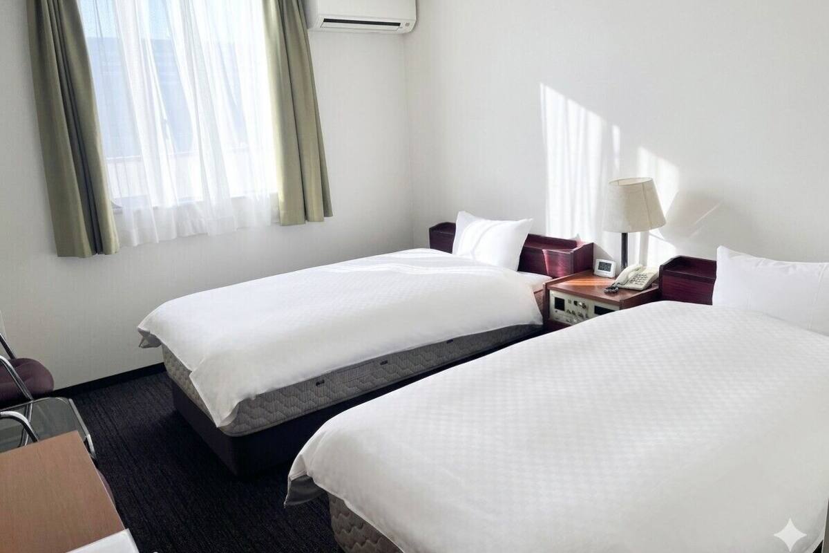 Une chambre d’hôtel avec deux lits, un bureau, une chaise, un appareil de climatisation et une fenêtre avec des rideaux.