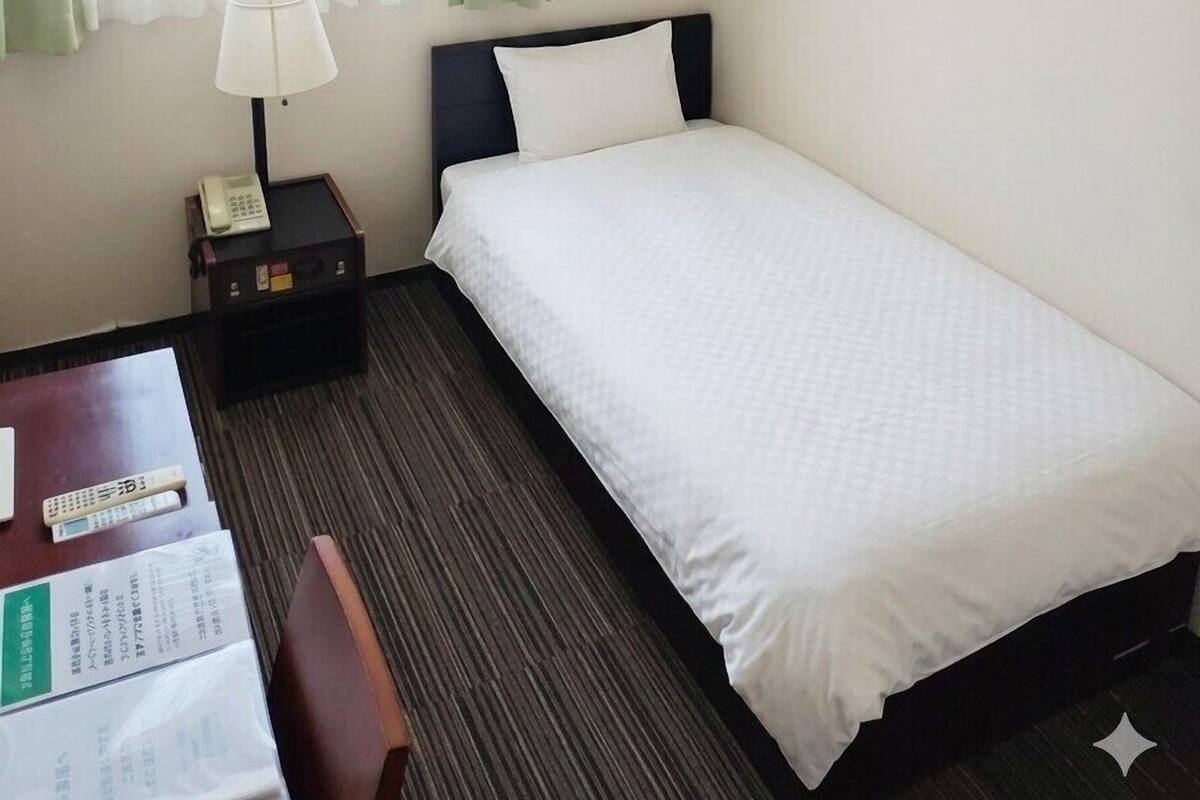 Une chambre d’hôtel avec un lit simple, une table de chevet avec une lampe, une chaise et un bureau avec un téléphone et des documents.