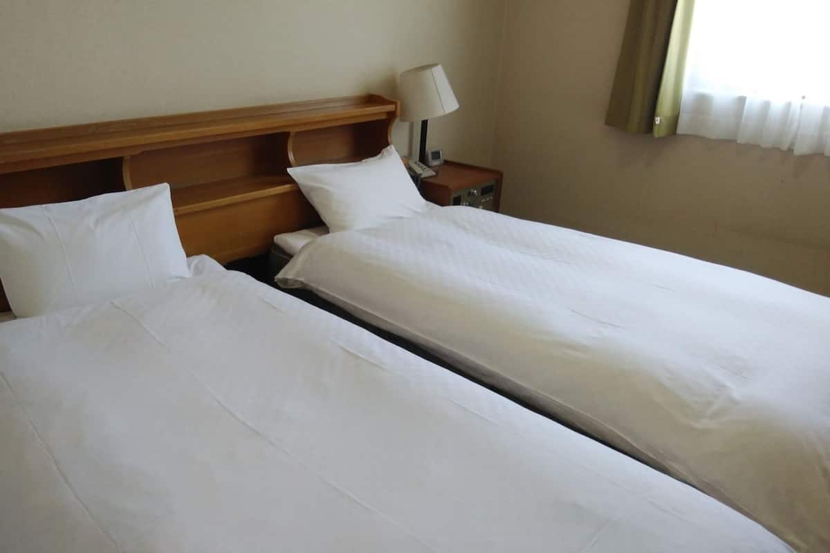 Une chambre d’hôtel avec deux lits simples, une tête de lit en bois, une table de chevet avec une lampe et une fenêtre avec des rideaux.