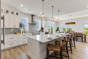 A spacious kitchen with white cabinetry, a large island, and a dining area with wicker chairs. - Serene Oasis | Luxury Oceanview Estate | Heated Pool | Game Room | Observation Deck | Sleeps 16 (Oak Island)