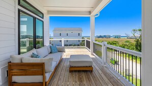 Une véranda couverte avec une terrasse en bois, un canapé blanc avec des coussins et une vue sur une maison et le paysage environnant.