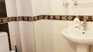 Egy fürdőszoba fehér csempékkel, fekete és barna színű, virágmintás díszítőcsíkkal, fehér WC-vel, adagolóval ellátott mosdókagylóval és a mosdókagyló felett elhelyezett tükörrel.