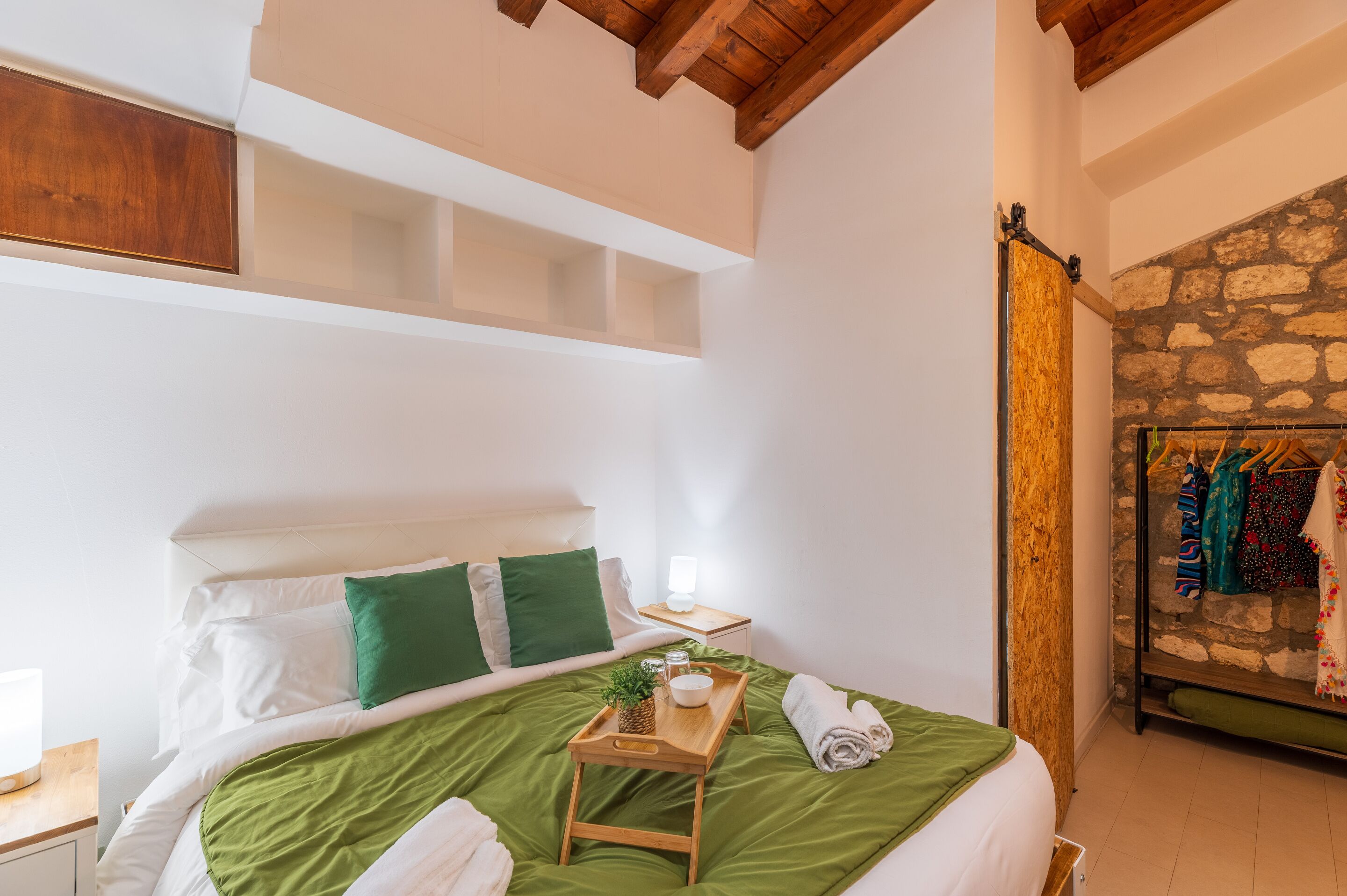 A bedroom with a bed, green pillows, a wooden tray with items, a nightstand, and a stone wall.