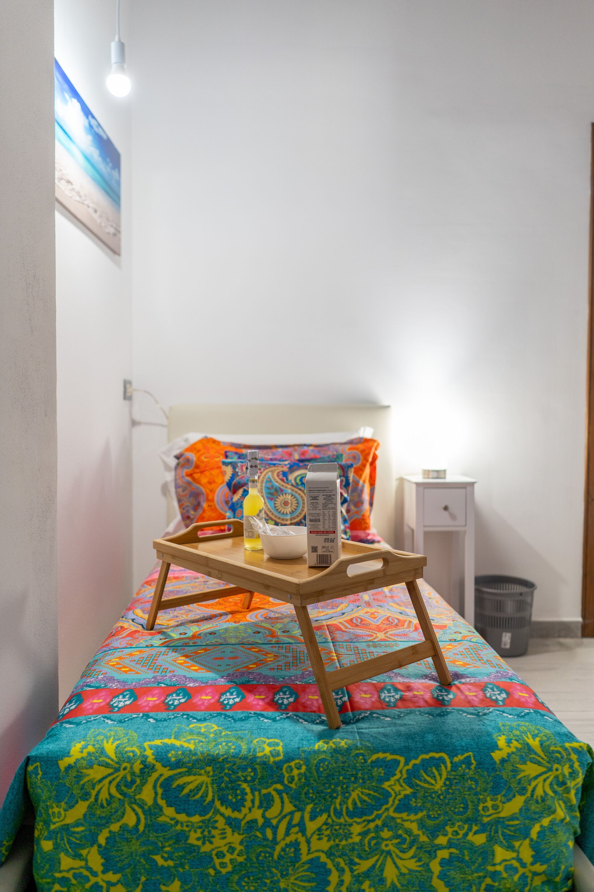 A small bed with a colorful bedspread, a bedside table with a lamp, and a wall-mounted picture.