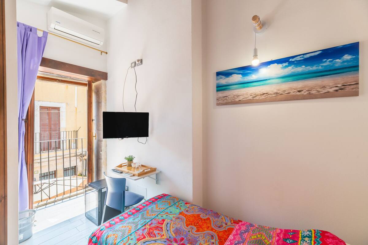 Een slaapkamer met een bed, een bureau, een stoel, een televisie en een schilderij van een strand.