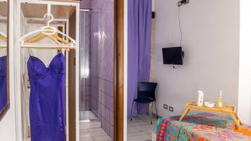 Un dormitorio con cama, una mesita de noche, un televisor montado en la pared, una silla y un baño visible a través de una puerta abierta.