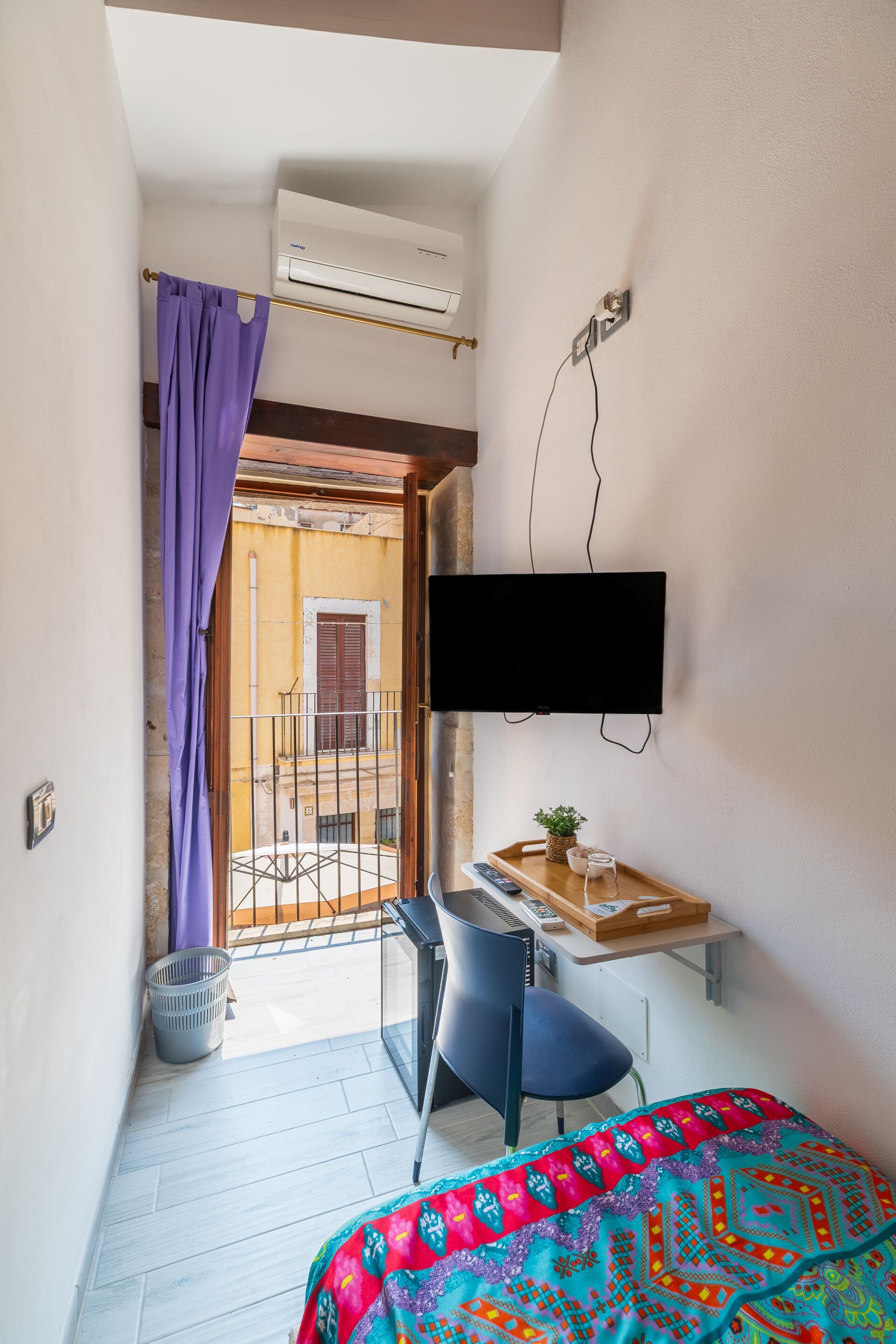 Een kleine, smalle kamer met een bed, een bureau met stoel, een aan de muur bevestigde televisie en een airconditioning.