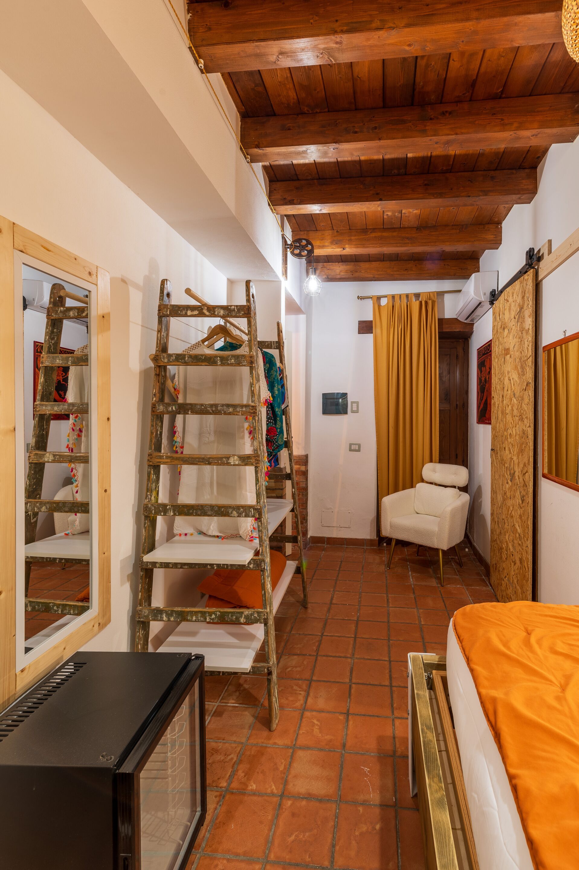 A compact room with a wooden ceiling, a bed with an orange blanket, a white chair, a wooden ladder, and a television.