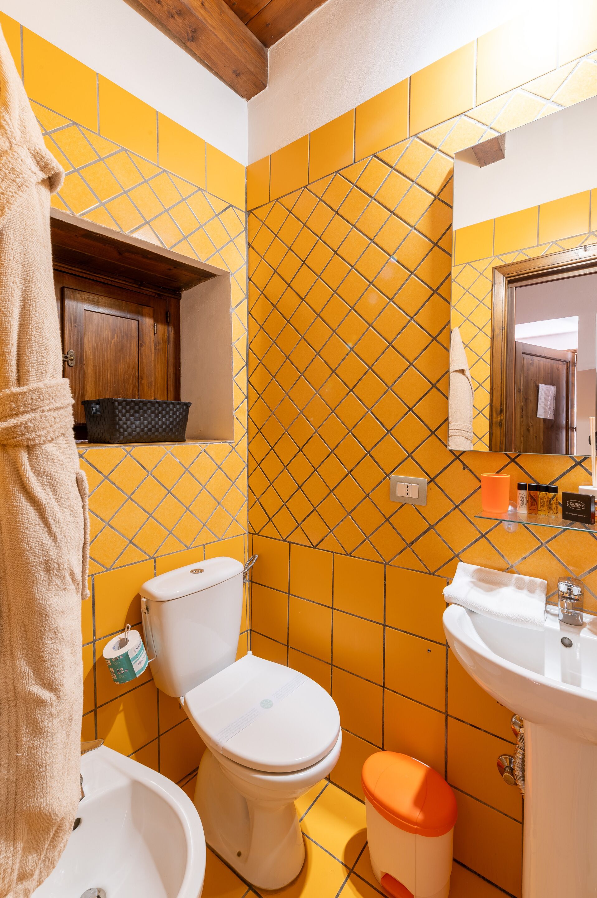 Un baño con baldosas amarillas estampadas, inodoro, bidé, lavabo y mueble de madera.