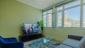 Un salón con televisión, una silla azul, un sofá gris, una mesa de centro de cristal y vistas a una obra en construcción a través de amplios ventanales.