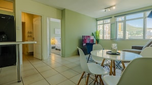 Una cocina moderna con refrigerador, un comedor con mesa y sillas, y vista a un paisaje urbano a través de amplios ventanales.