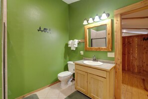 Un baño con pared verde, tocador de madera, inodoro blanco y espejo con luces.