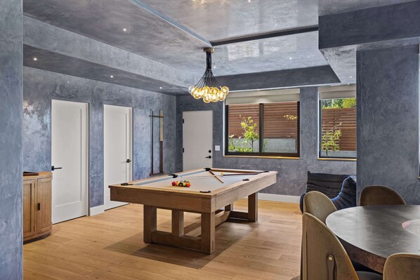 Una mesa de billar moderna en un espacio amplio con piso de madera, una zona de comedor con mesa redonda y un gran ventanal con vistas a zonas verdes.