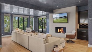 A modern living room with a sectional sofa, a fireplace, a TV mounted on the wall, and large windows offering a view of the outdoors.