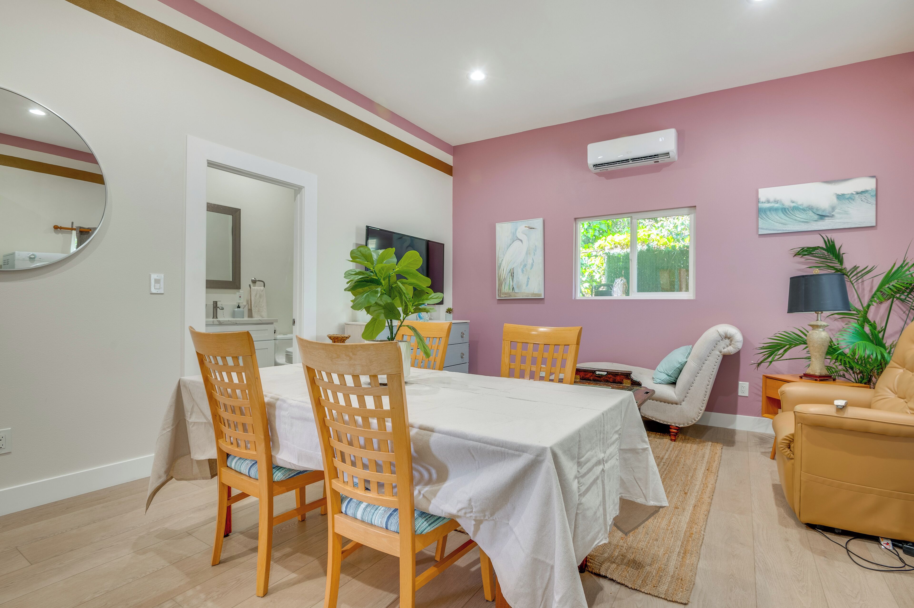 A dining area with a table set for four, surrounded by wooden chairs. There is a potted plant on the table, and a white armchair is visible in the background.