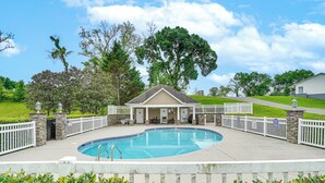 Un área de piscina con zona de estar cubierta, pilares de piedra y una valla blanca que rodea la piscina.