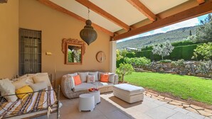 A covered outdoor seating area with a sofa, ottoman, and round tables, overlooking a landscaped garden and hills. - Alondra - Javea, Costa Blanca (Javea)