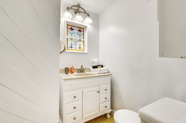 Une salle de bain avec un meuble-lavabo blanc, un lavabo avec des articles de toilette, des toilettes et un vitrail au-dessus du miroir.