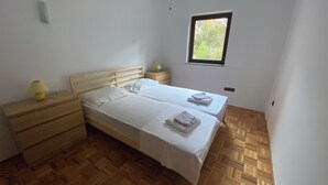 Un dormitorio con dos camas individuales, cabeceros de madera, una mesita de noche con lámpara, una ventana con vistas a zonas verdes y un suelo de parquet de madera.