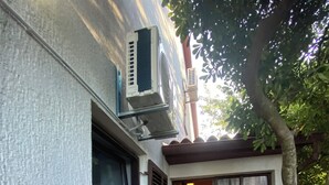 Un edificio de fachada blanca, una ventana con contraventanas oscuras, un pequeño porche y un árbol en primer plano.