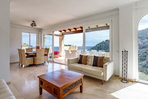 Un soggiorno spazioso con un tavolino in legno, un divano beige con cuscini marroni, poltrone in vimini e un balcone vista mare.