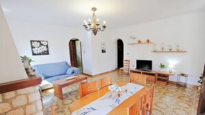A living room with a sofa, dining table, and chairs. There is a TV and a chandelier.