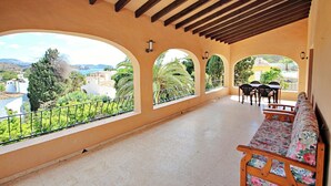 A covered balcony with a sofa and chairs, large arched windows, and a view of greenery and buildings.