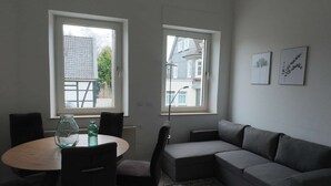 Ett modernt vardagsrum med en grå soffa, ett runt bord med stolar och två fönster med utsikt över byggnader och träd.