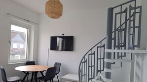 A modern room with a round wooden table, black chairs, a woven pendant light, a flat-screen TV, and a spiral staircase. - Duplex apartment with two bedrooms and a living room (Solingen)