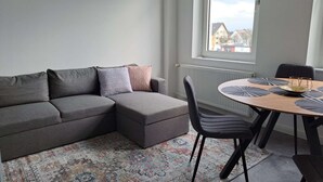 A modern living room with a grey sectional sofa, a round wooden dining table with black chairs, and a patterned rug. - Duplex apartment with two bedrooms and a living room (Solingen)