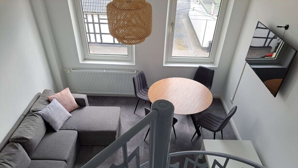 A modern living room with a grey sectional sofa, a round table, and two black chairs. There are two windows with a view of the street, and a hanging woven light fixture. - Duplex apartment with two bedrooms and a living room (Solingen)