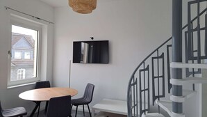 A modern interior with a round wooden table, black chairs, a woven pendant light, a black-framed mirror, and a grey spiral staircase. - Duplex apartment with two bedrooms and a living room (Solingen)
