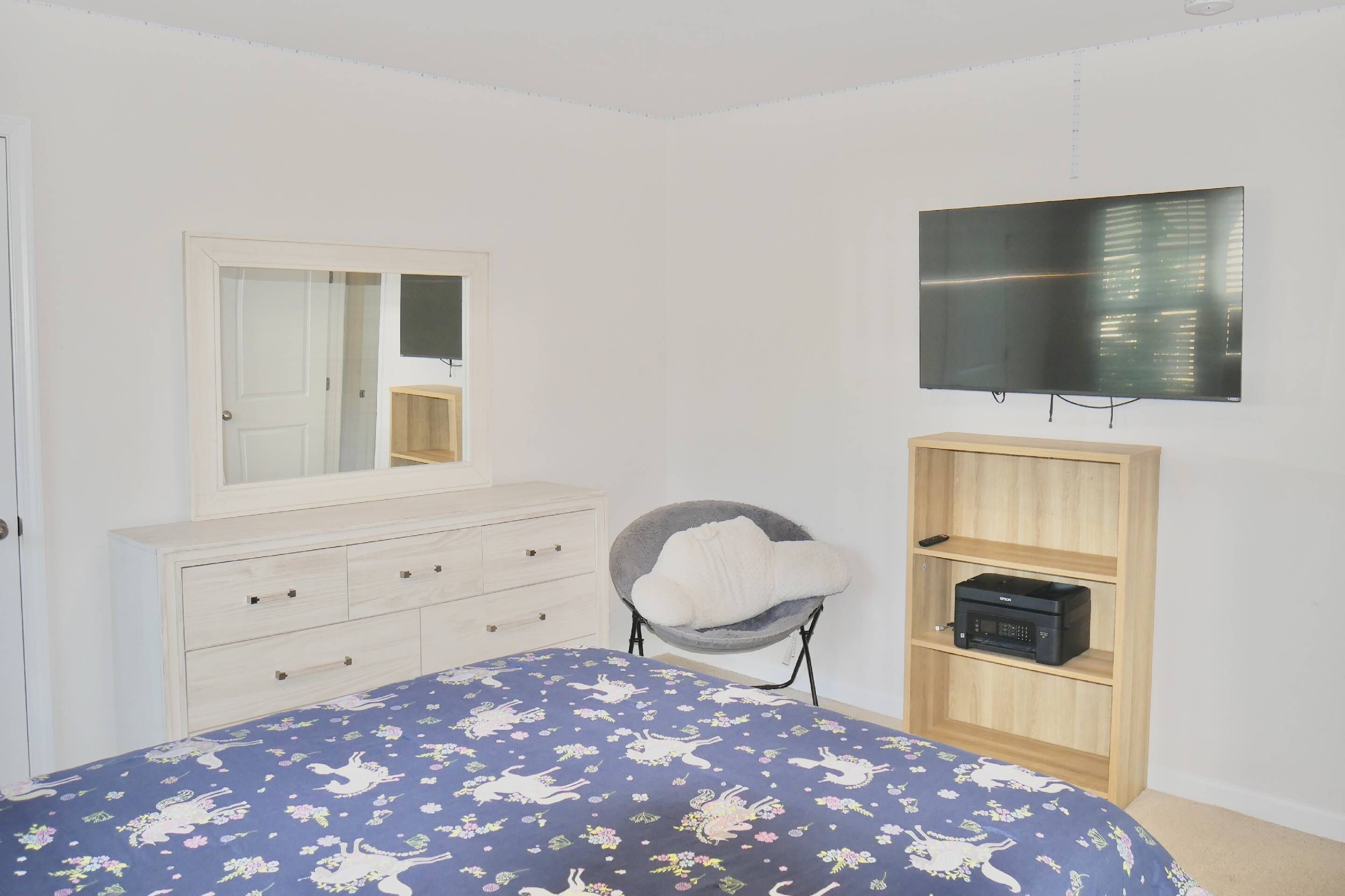 Un dormitorio con cama, cómoda, silla, televisor y estante con un dispositivo.