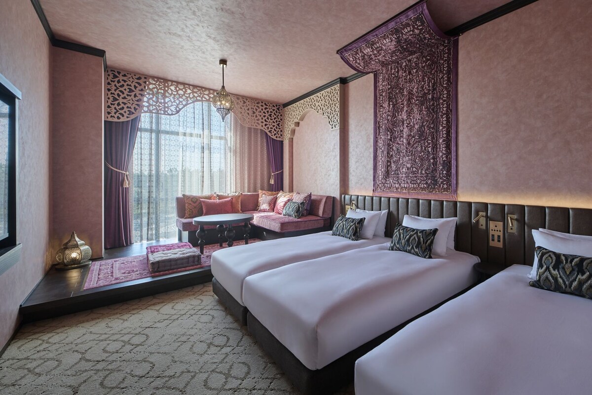 A hotel room with two beds, a sofa, a chandelier, and a large window with curtains.