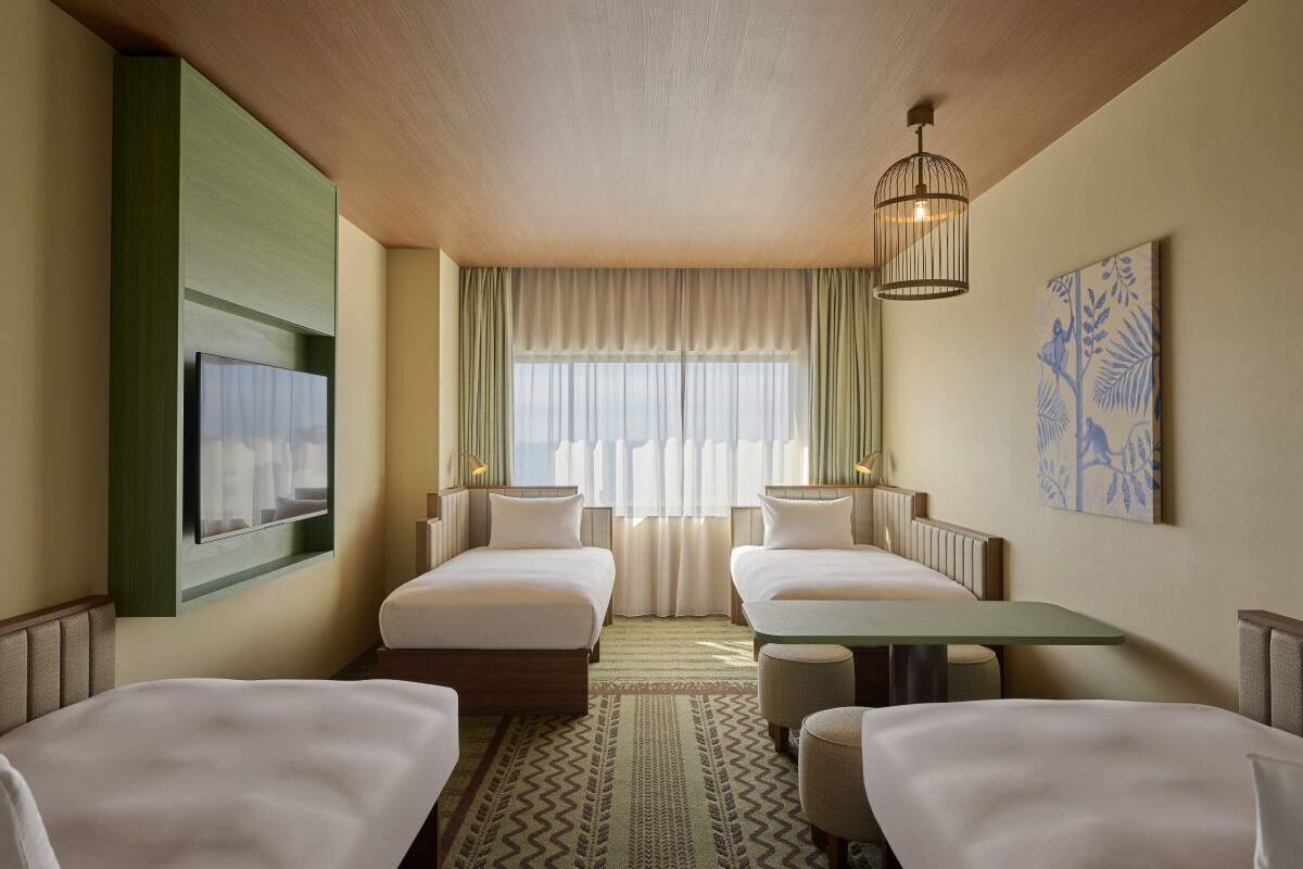 A hotel room with two beds, a dining table, a large window with curtains, a hanging light fixture, and a wall-mounted artwork.