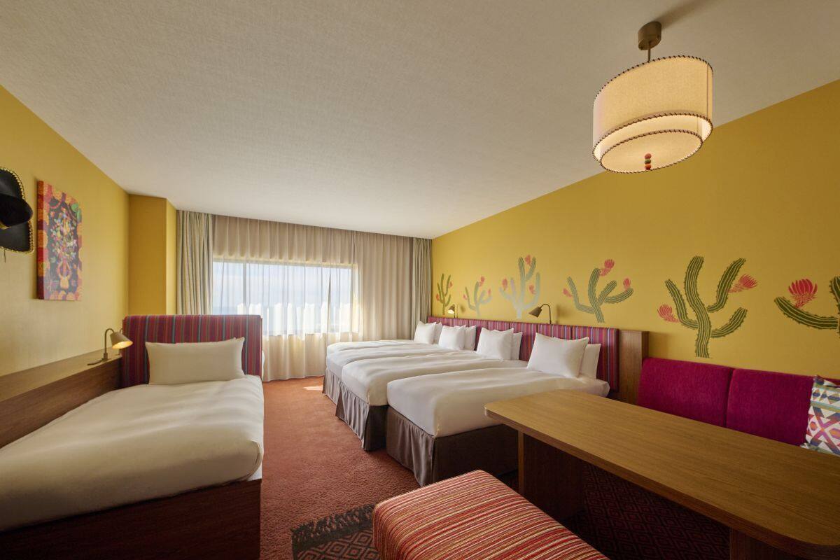 A hotel room with three beds, a dining table, and a large window with curtains.