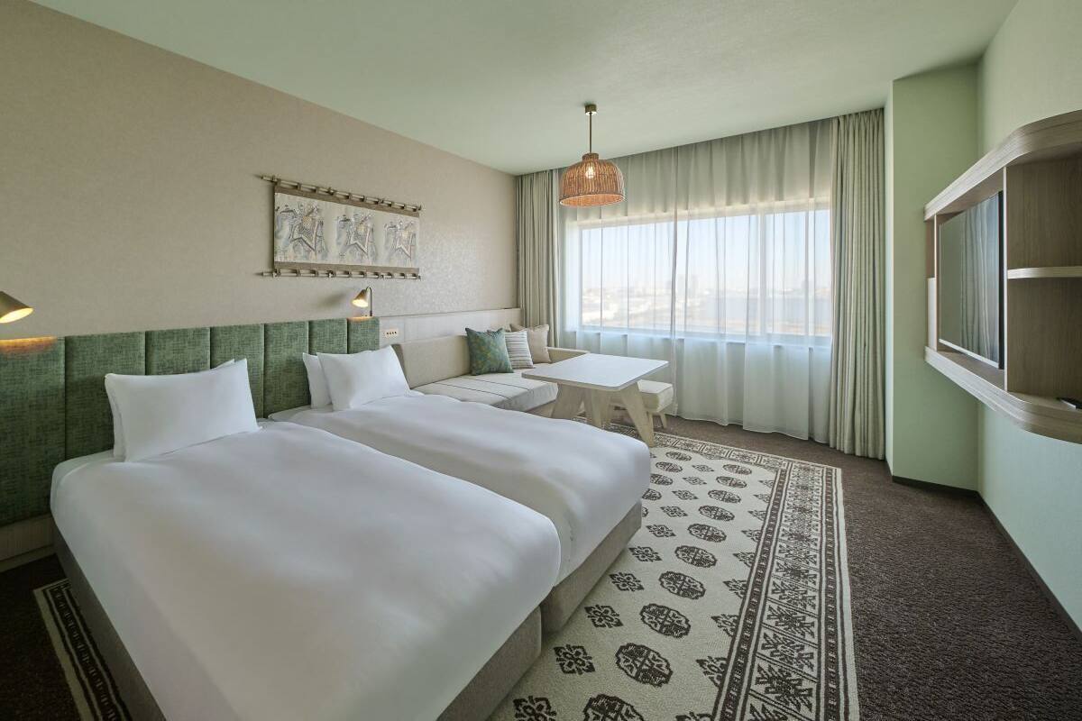 A hotel room with two beds, a sofa, a small table, a patterned rug, and a large window with curtains.