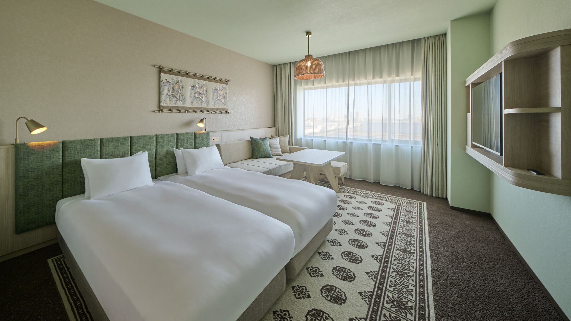A hotel room with two beds, a small table, a sofa, a patterned rug, a window with curtains, and a wall-mounted artwork.