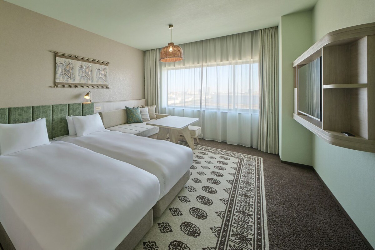 A hotel room with two beds, a small table, a sofa, a patterned rug, a window with curtains, and a wall-mounted artwork.