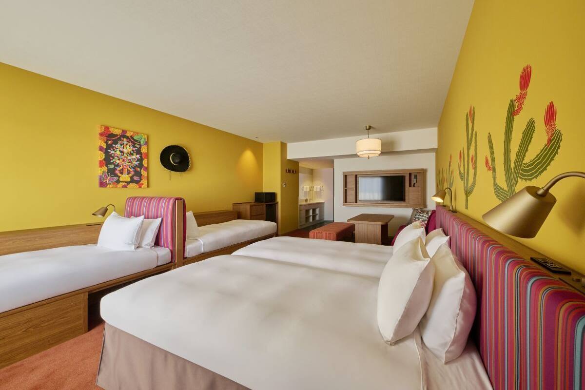 A hotel room with two single beds, a sofa, a TV, and a cactus mural.