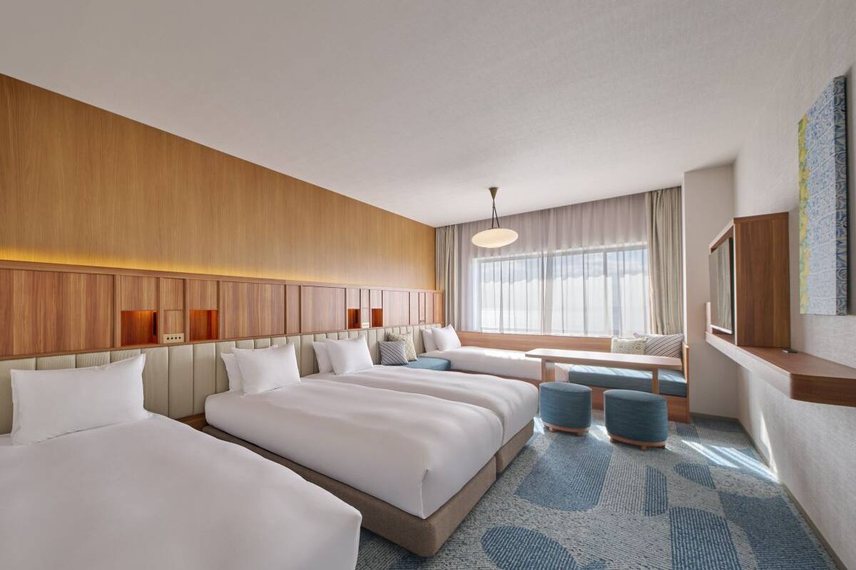 A hotel room with three beds, a seating area, a large window with curtains, and a wall-mounted TV.