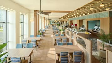 A spacious hotel dining area with long tables, blue patterned chairs, and a bar area with a view.