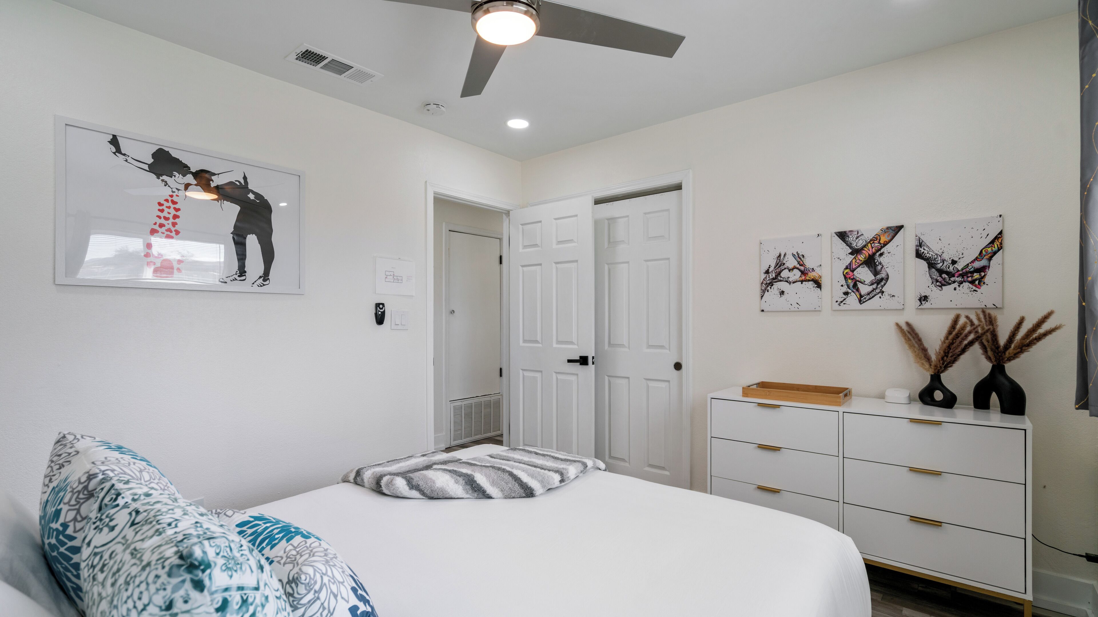 A bedroom with a bed, a ceiling fan, a dresser, and artwork on the walls.