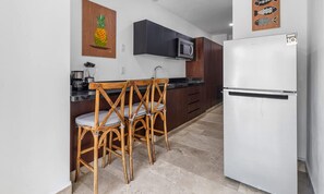 Una cocina con un refrigerador blanco, sillas de madera, un microondas y un cuadro con piña en la pared.