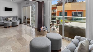 A modern living room with a grey sectional sofa, grey ottomans, a flat-screen TV, and a glass door leading to an outdoor balcony with a table and chairs. - Risco 305: Downtown, Rooftop Pool, & Beach Access (Bucerías)